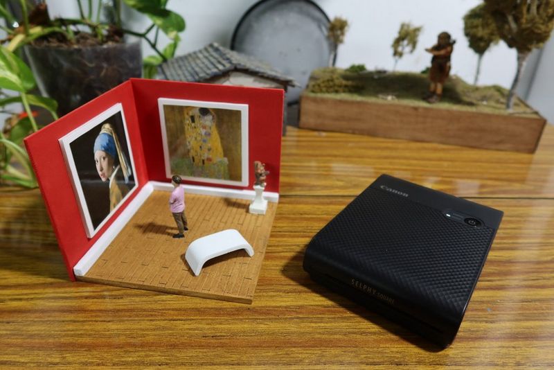 A mini art gallery diorama is positioned on a tabletop next to a Canon SELPHY Square QX10 printer.