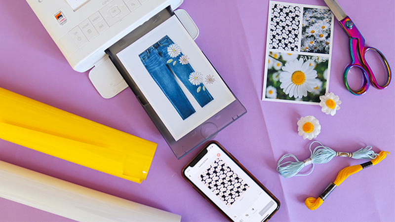 White SELPHY printer with a picture of jean trousers with daisies being printed. A phone showing a daisy print on the screen and a picture of different images of daisies in the corner.