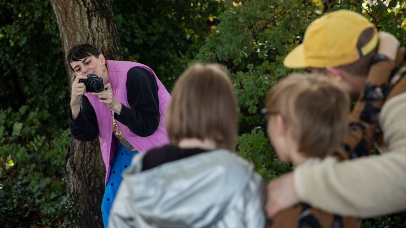 In a forest setting, a woman holding a Canon EOS R100 leans over to capture a family photo of two children and a man wearing a yellow cap.