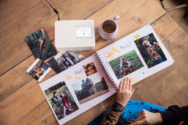 A photo album lies open on a wooden desktop, with some loose prints and a Canon SELPHY printer.