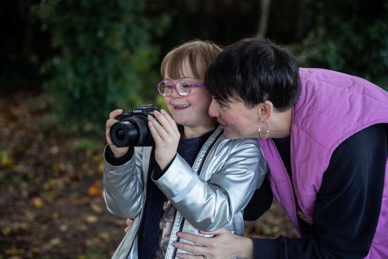 In a forest setting, a young girl holds a Canon EOS R100 while her mother leans over her shoulder and talks to her.
