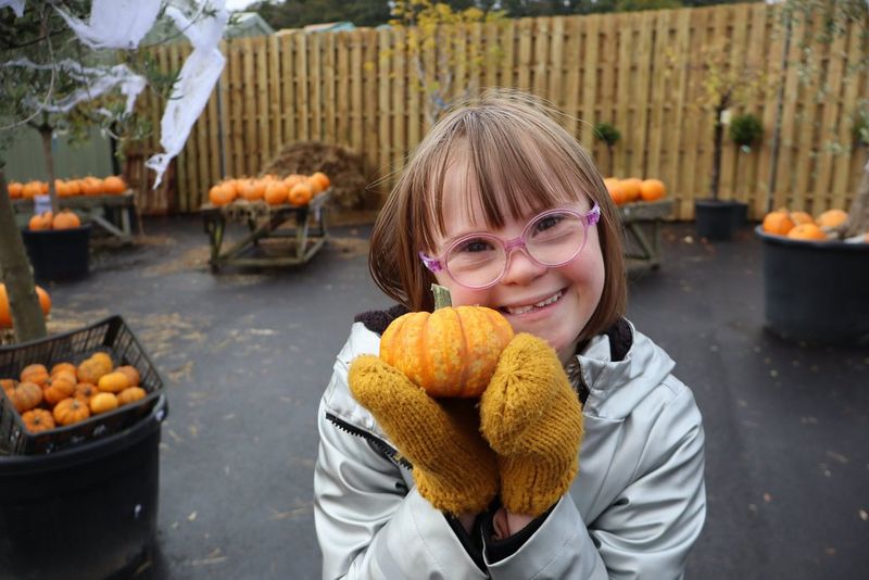 In a photo taken with a Canon EOS R100, a girl wearing mittens and a coat holds a small pumpkin up to the camera, with stands of pumpkins out of focus in the background.
