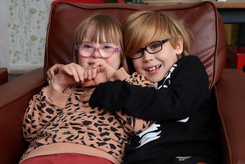 In a photo taken with a Canon EOS R100, two smiling children playfully hug while sitting together on an armchair.