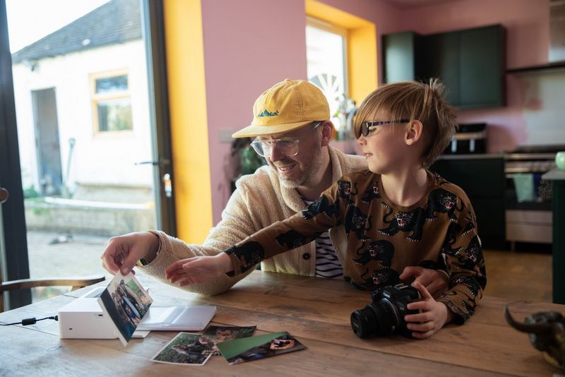 A father and son sit at a wooden table, the son with one hand on a Canon EOS R100 camera on the table, and the two looking at prints that have emerged from a Canon SELPHY printer. 