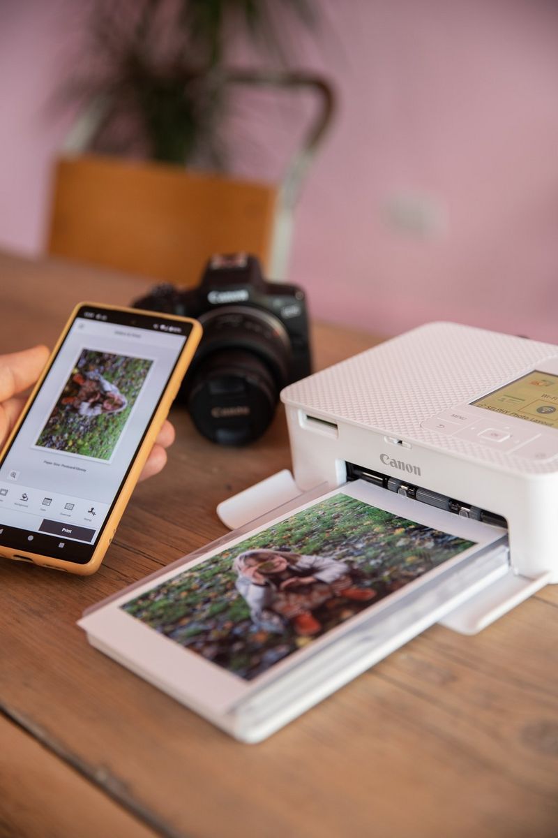 A photo of a child surrounded by leaves is displayed on a smartphone as a print of the image emerges from a Canon SELPHY printer beside it, with a Canon EOS R100 camera in the background.