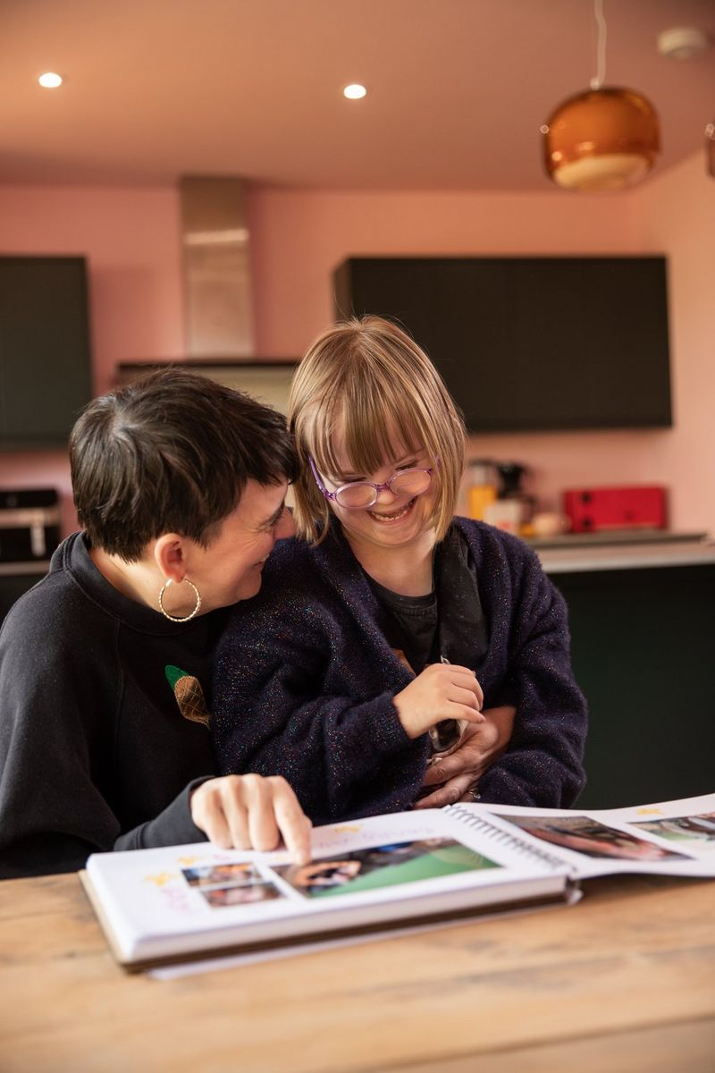 A mother and daughter sit at a table, smiling and laughing together as they look through a photo album.