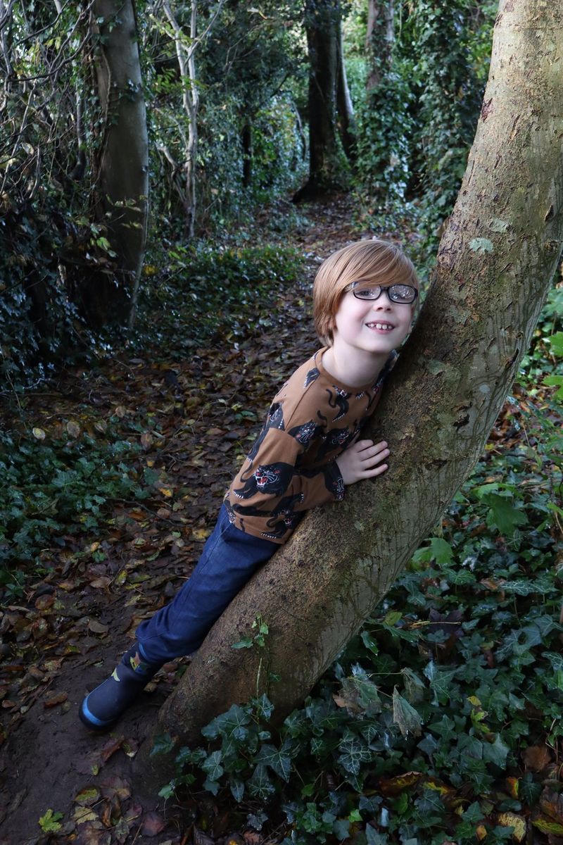 In a photo taken on a Canon EOS R100, a child leans against a tree at a tilted angle against a forest background.