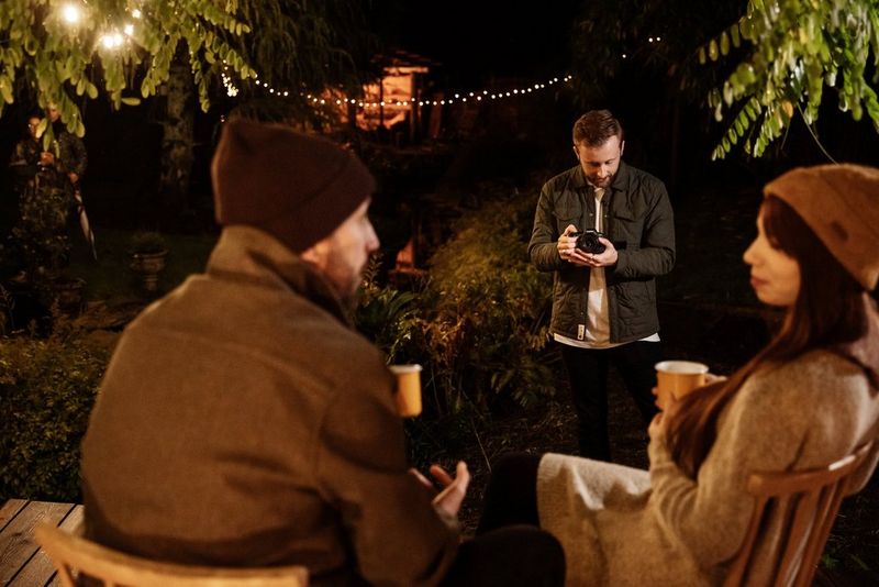 A couple chat while sitting and holding hot drinks, and are being filmed by a man holding a Canon EOS R6 Mark II.