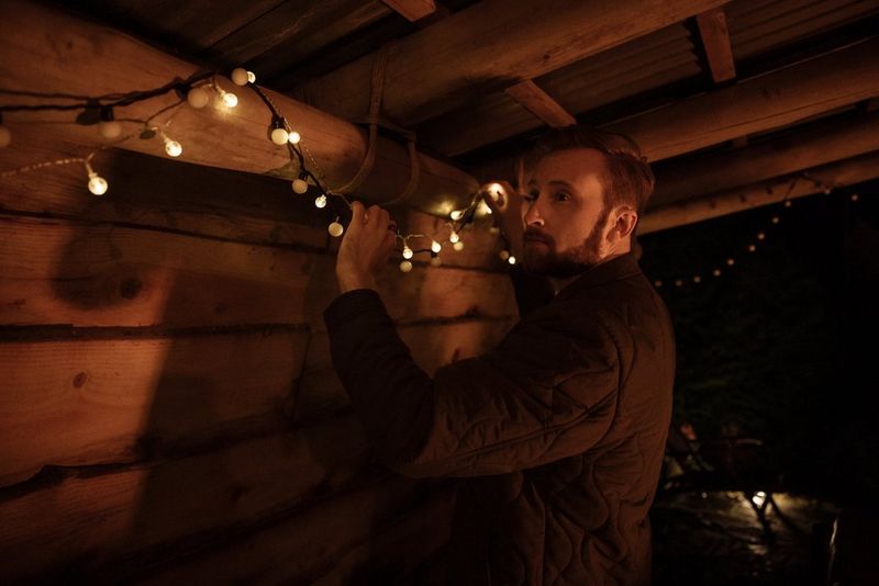 A man hangs a string of fairy lights on a large wooden beam. 