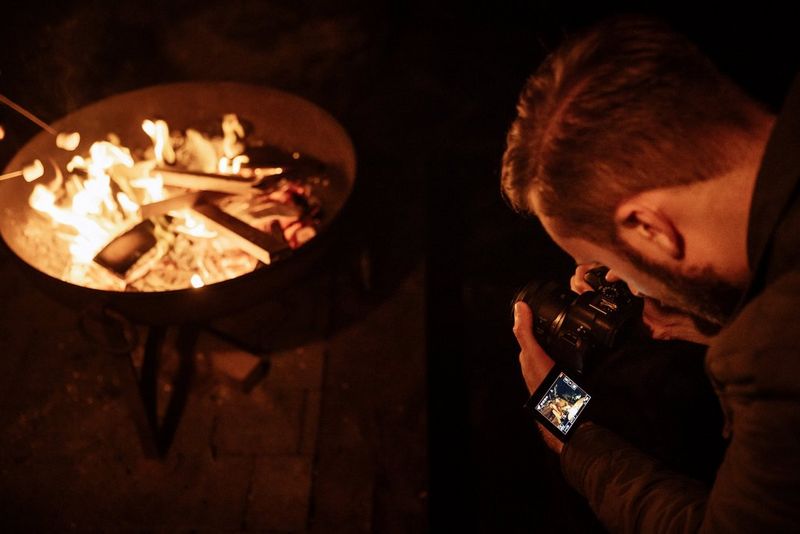 A man leans down with a Canon EOS R6 Mark II camera, shooting across the top of a campfire. A couple sitting beside the fire can be seen on the camera touchscreen.