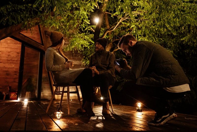 A photographer crouches down to film a young couple in winter clothing as they sit chatting on a wooden deck. The scene is lit by small lamps. 