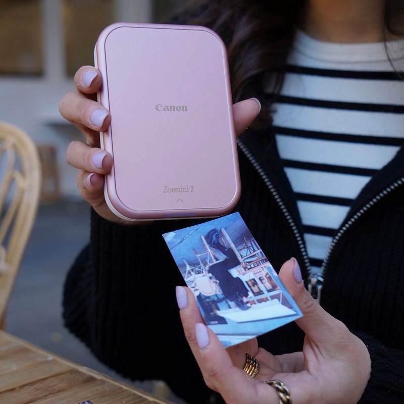 A woman sat at an outdoor table holds up a Canon Zoemini 2 printer and a just-printed photograph.