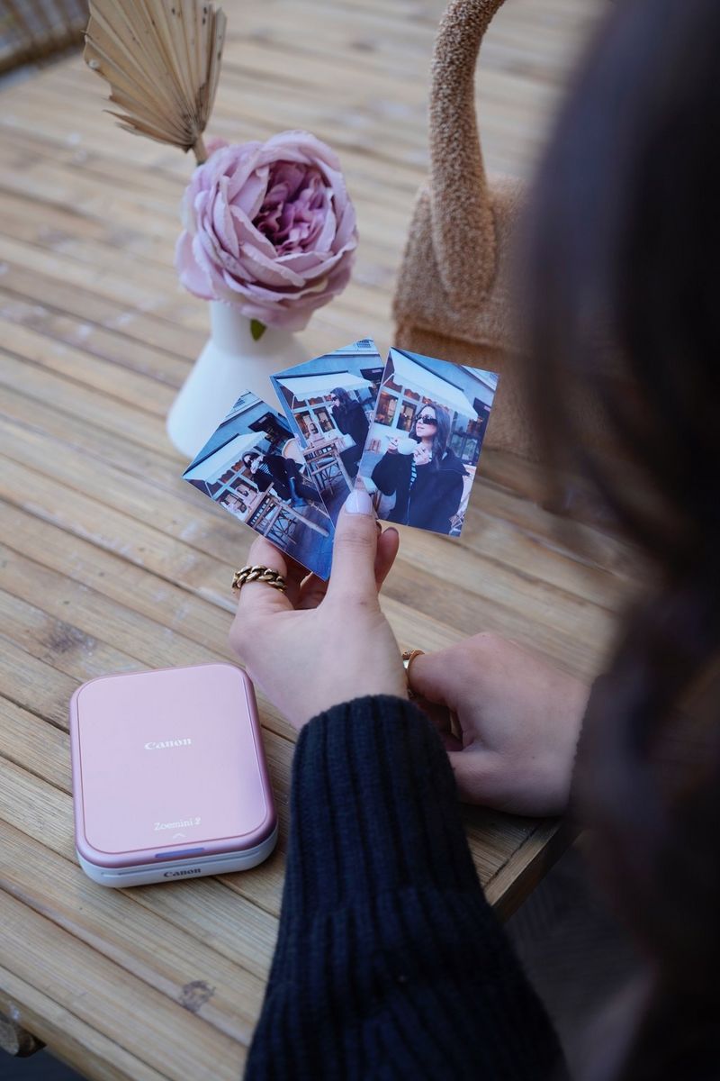 A woman sitting at an outdoor table holds up a selection of small photographs. On the table is a Canon Zoemini 2 printer and a pink rose in a vase.