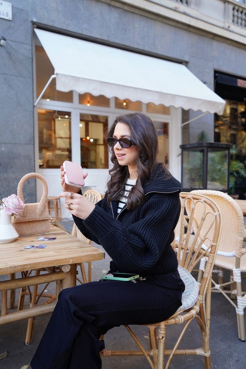 A woman with long dark hair and a smart black outfit sits at an outdoor cafe table, holding up a Canon Zoemini 2 printer.