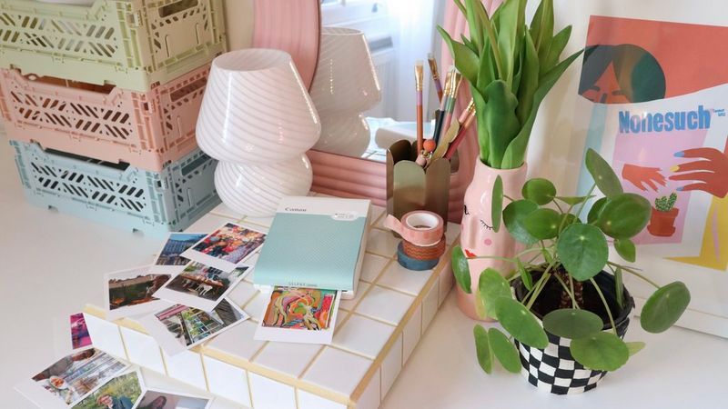 A top-down image of a Canon SELPHY Square QX10 printer on a desk surrounded by mini prints, stationery, a small lamp, a vase of flowers and a plant. 