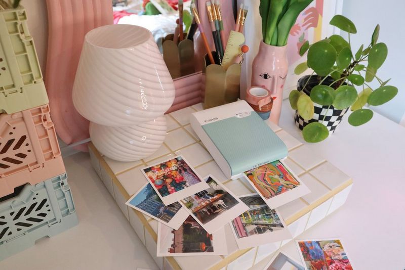 A green Canon SELPHY Square QX10 prints a colourful image. On the desk around it are several more prints, as well as a pen pot, flowers, three colourful boxes and a potted plant. 