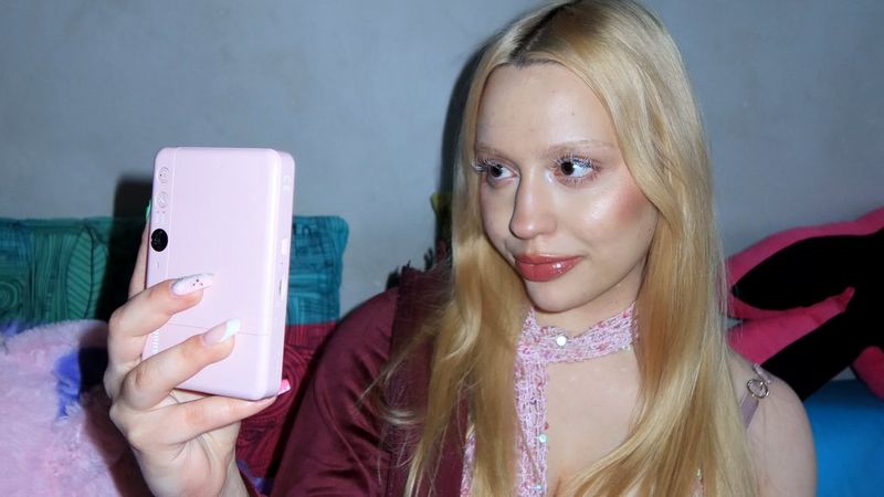 Style influencer Molly Wójcik, wearing a burgundy jacket and a sequinned scarf, holds up a rose gold Canon Zoemini S2 to take a selfie.