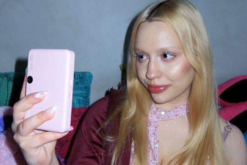 L'influencer di stile Molly Wójcik, con una giacca bordeaux e una sciarpa di paillettes, solleva una stampante Canon Zoemini S2 oro rosa per scattare un selfie.