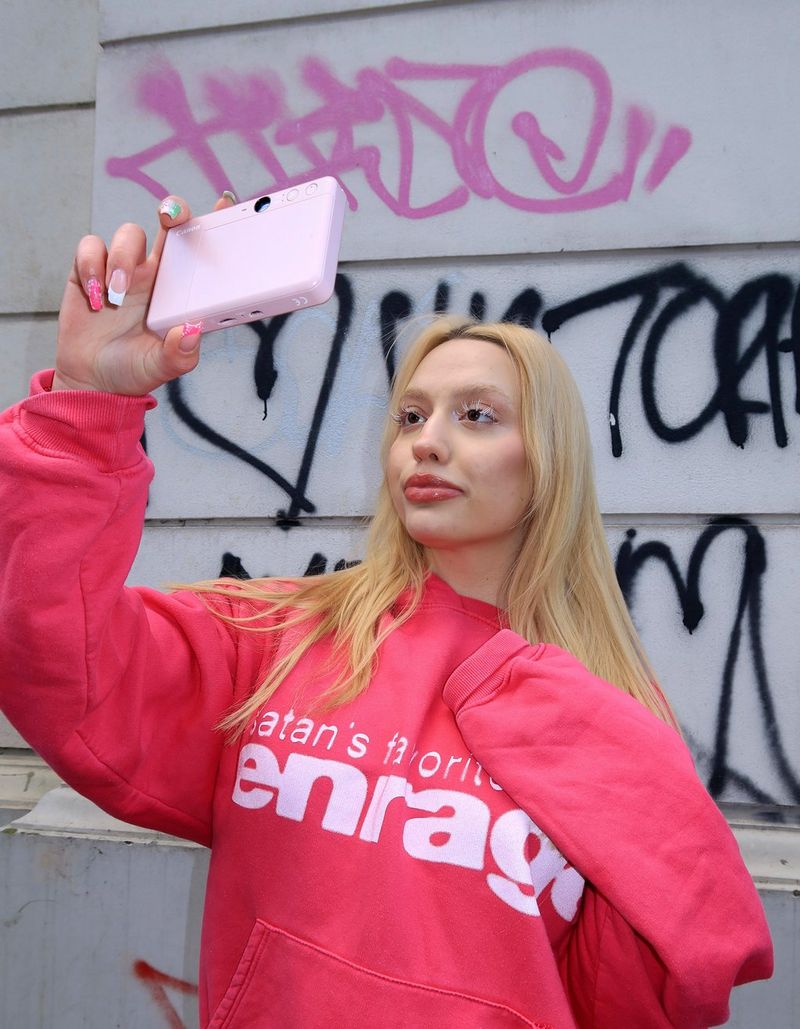 Style influencer Molly Wójcik wearing a pink sweatshirt and posing for a selfie while holding a Canon Zoemini S2. 