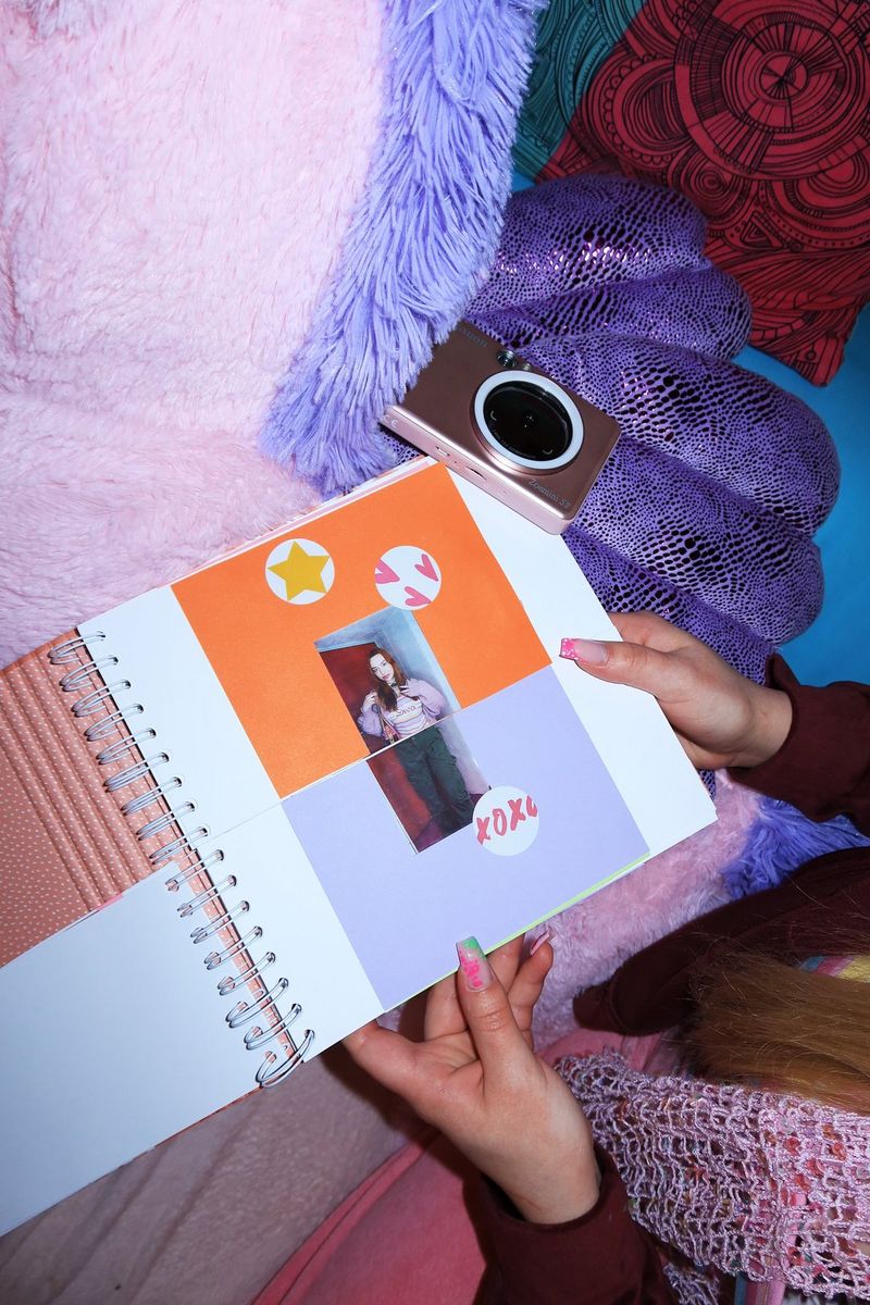 A pair of hands holds open a fashion lookbook with outfits cut in half to enable the top and bottom to be turned independently. A Canon Zoemini S2 is on a pile of cushions beside the book. 