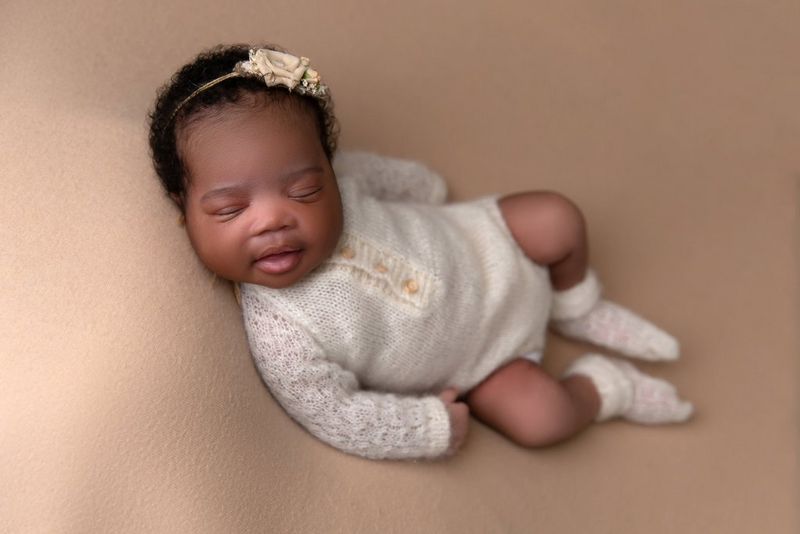 A black baby girl wearing off-white woollen onesie is smiling in her sleep.
