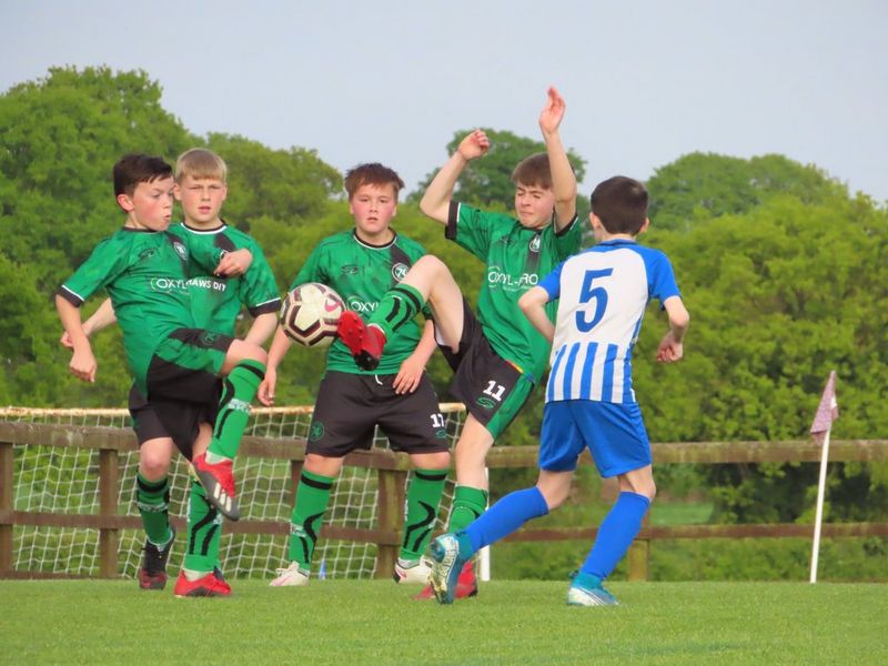 Boys playing football, four in green kit, one with his leg raised in the air to kick to ball, and one player in blue.