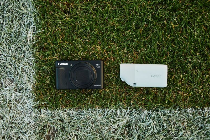A Canon PowerShot SX740 HS and Canon PowerShot ZOOM placed on the grass of a playing field, within the white, painted lines.
