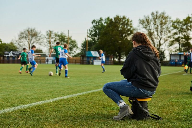 Molly Darlington sitting on a small stool by the side of a football pitch, watching a game taking place.