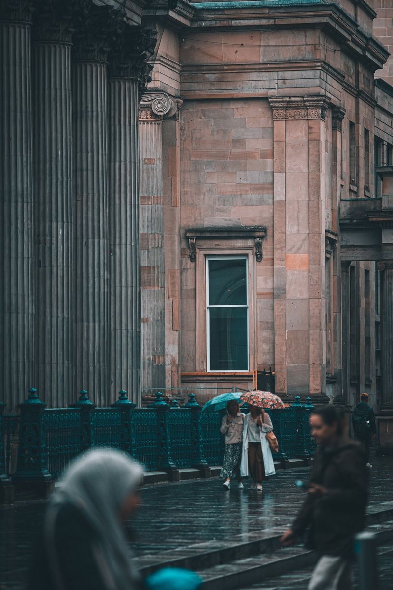 Two figures holding umbrellas walk in front of a large, imposing building. Taken by street photographer Andres McNeill. 