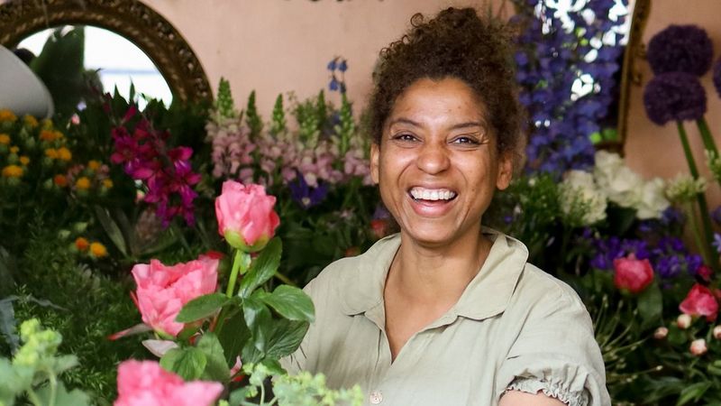 A smiling florist in a pale green dress surrounded by colourful flowers.