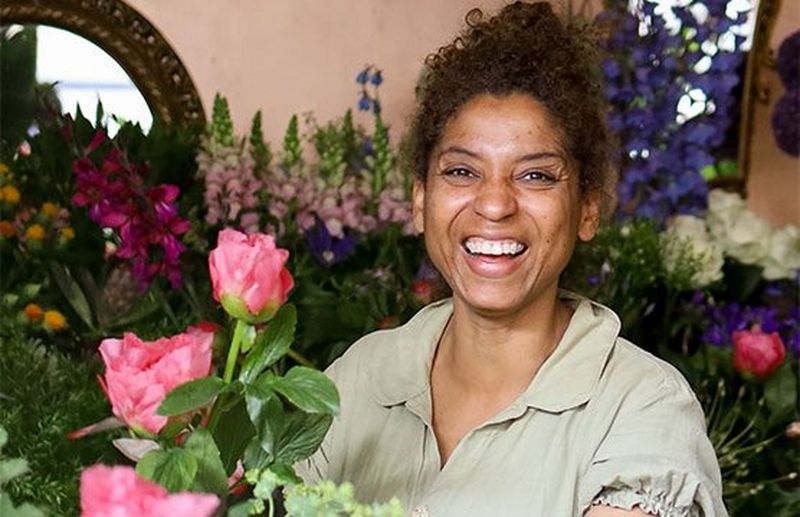A smiling florist in a pale green dress surrounded by colourful flowers.