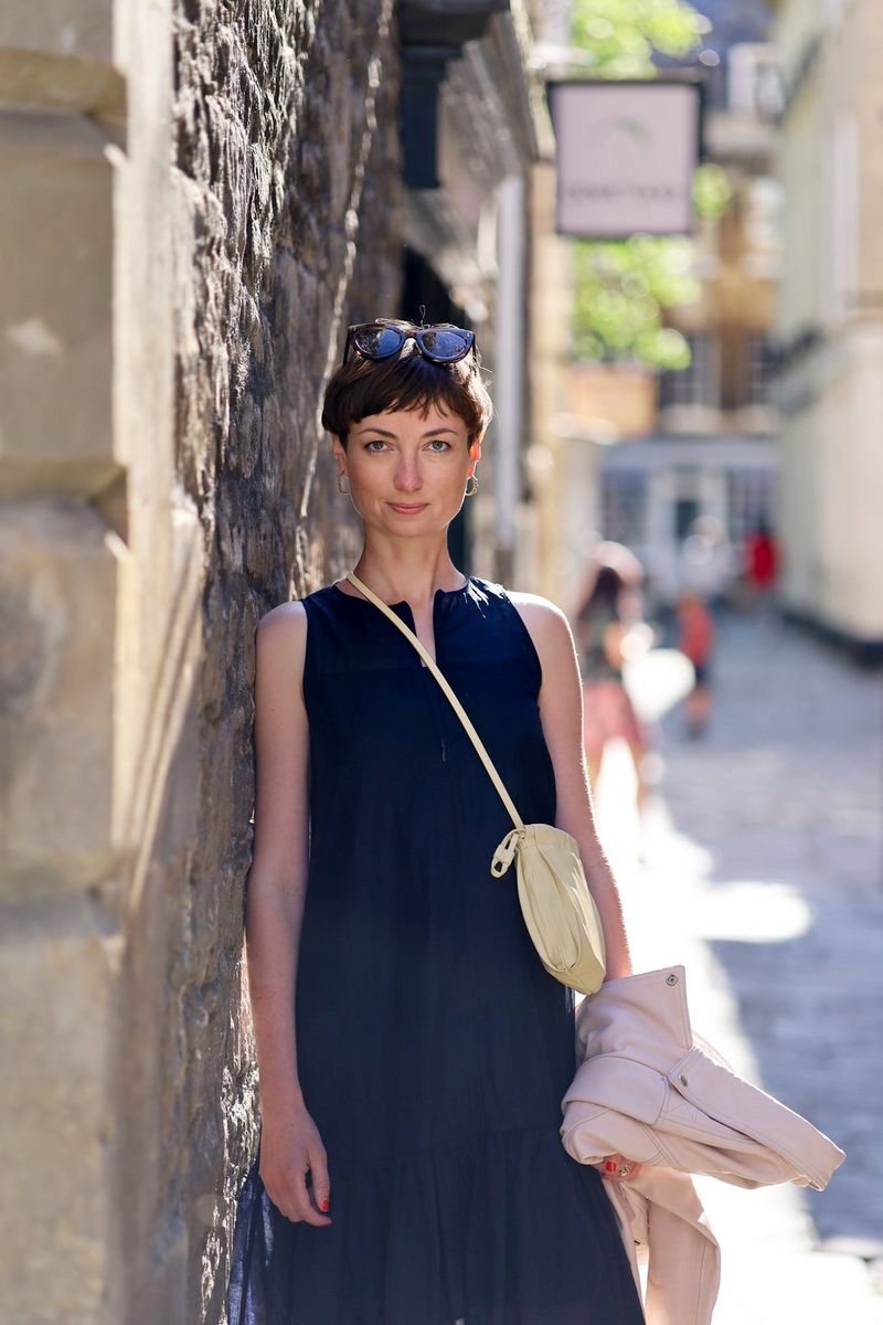 A woman in a blue sleeveless dress and with sunglasses on her head leaning against a stone wall. 