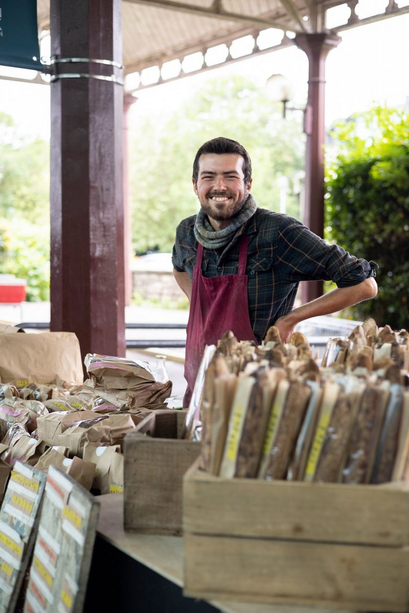 A smiling stall holder, wearing a scarf and a burgundy apron, standing behind his wares.