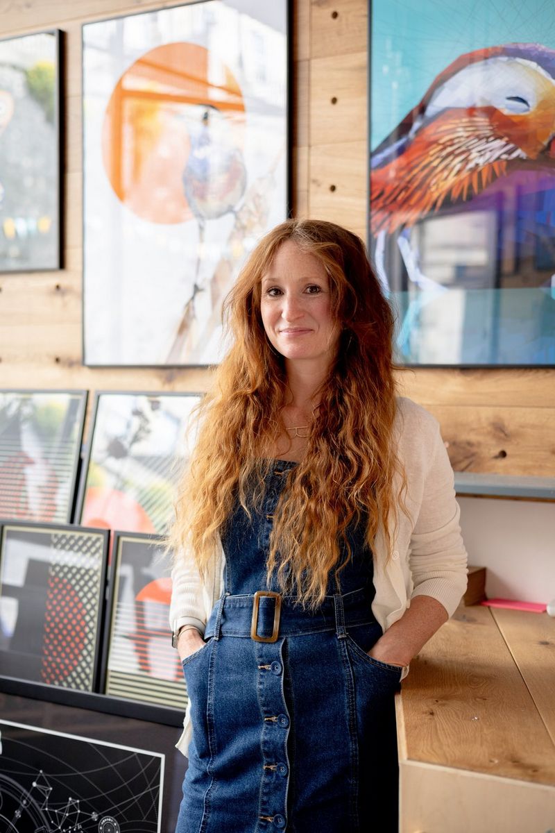 A woman with ginger hair in a denim dungaree dress standing in front of paintings hanging on a wood-panelled wall. 