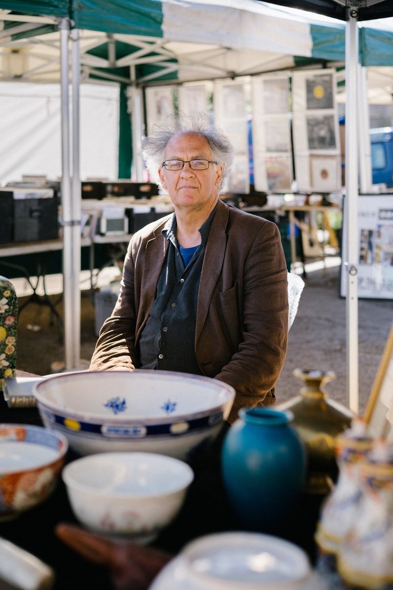 A grey-haired man in a dark brown jacket sitting behind a market stall selling antique bowls and vases.