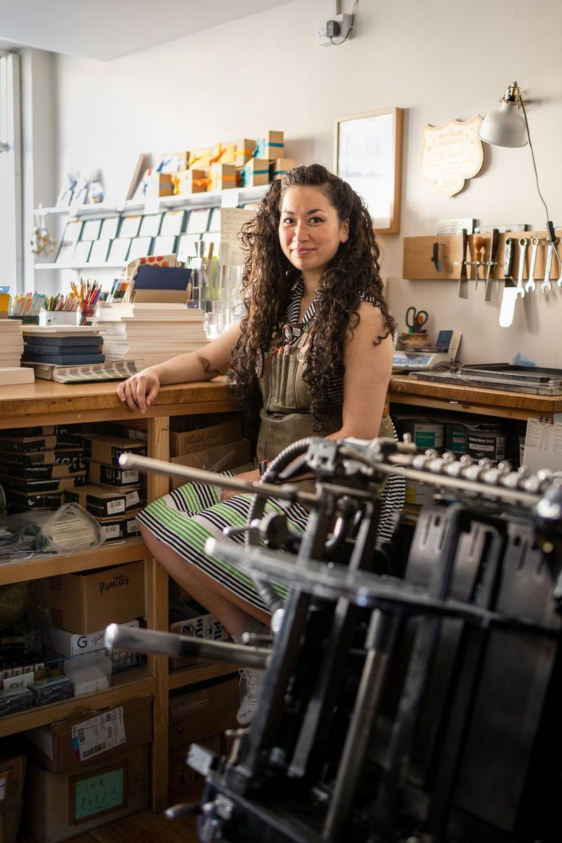 A woman with long, curly dark brown hair sits at a shop counter next to a letterpress machine.