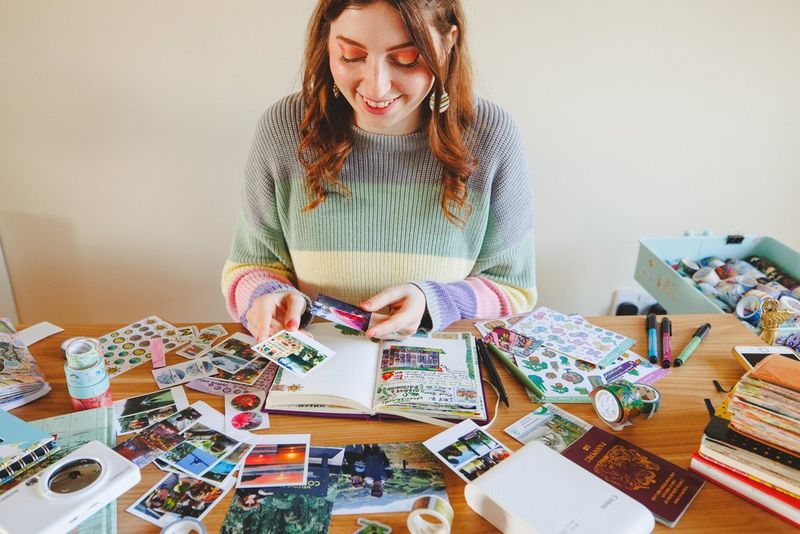 A woman with long dark hair and wearing a pastel striped jumper sits at a desk choosing photos to go in a travel journal. The journal is surrounded by photos, stickers, washi tape, a Canon Zoemini S2, a Canon SELPHY Square QX10 with a photo emerging from it and more.