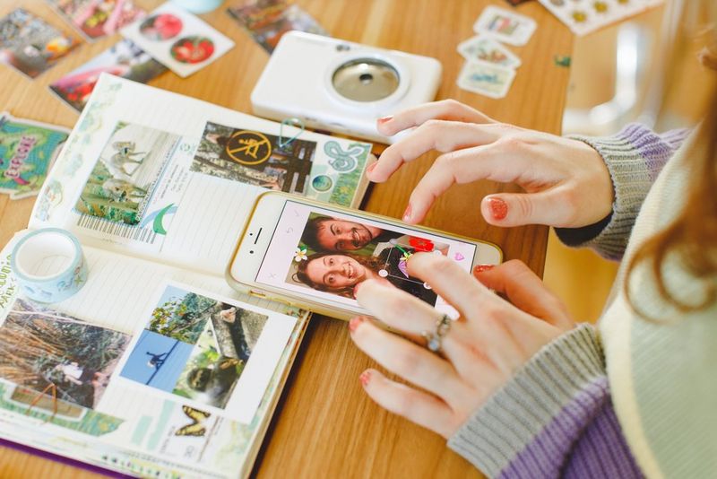 Hands tap at a smartphone showing a photograph with graphics being added. The phone is lying on an open travel journal, and a Canon Zoemini S2 lies to the side.