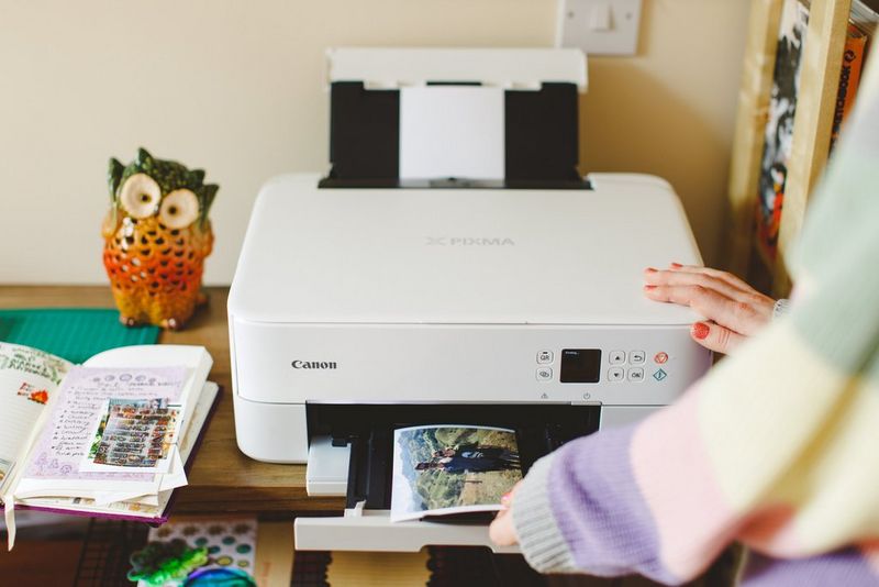 A woman just out of shot stands by a Canon PIXMA printer on a small table, removing a photograph that has just been printed.