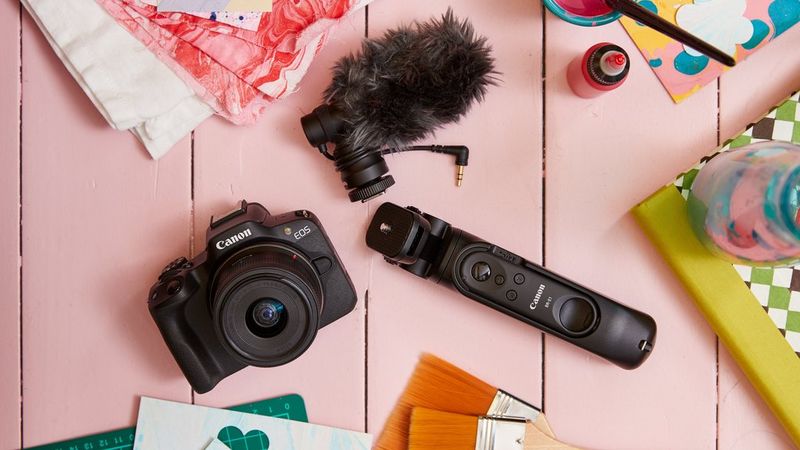 A flatlay of a Canon EOS R50 camera, external microphone and tripod grip on a pink wooden background, surrounded by crafting materials.