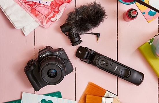 A flatlay of a Canon EOS R50 camera, external microphone and tripod grip on a pink wooden background, surrounded by crafting materials.