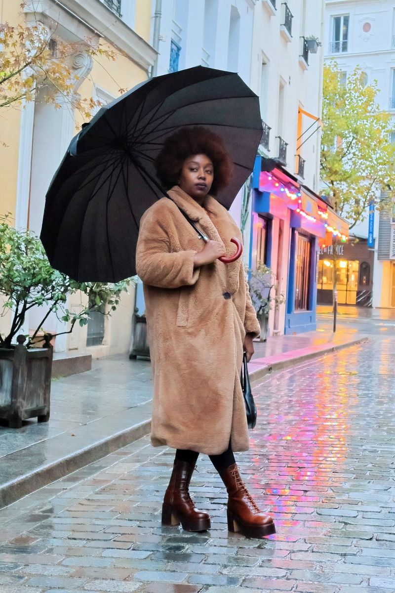A woman in a brown fluffy coat stands holding an umbrella in a cobbled street alongside illuminated shopfronts. Taken on a Canon EOS R50.