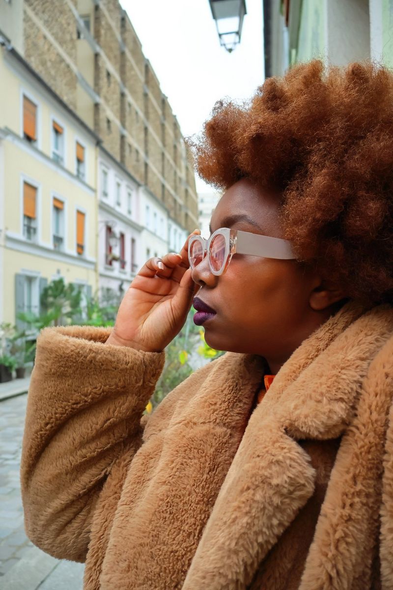 A side profile of a woman in a brown fluffy coat holding her hand to her glasses. Taken on a Canon EOS R50.