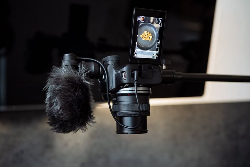A Canon EOS R50 camera with a microphone attached is held by a vertical stand with the lens facing down. A frying pan is displayed on the camera's vari-angle touchscreen.