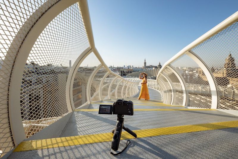 A Canon EOS R10 positioned on a tripod between curved railings to film a woman in the distance.