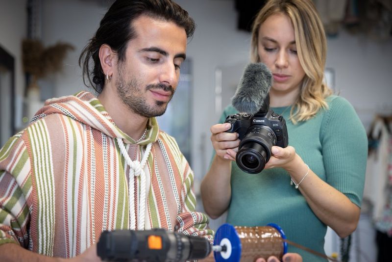 A woman, holding a Canon camera with a Stereo Microphone DM-E1 attached, filming a man with a drill.