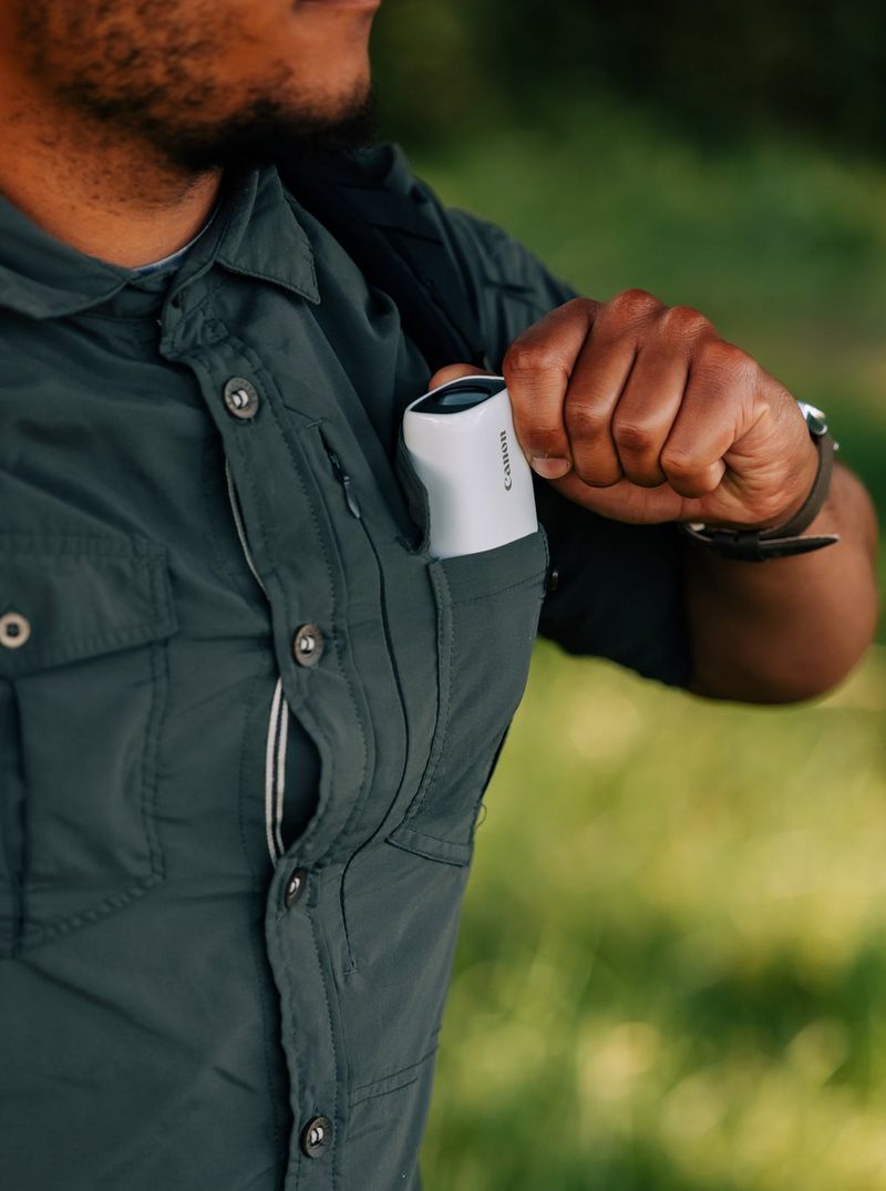 A man in the woods putting a Canon PowerShot ZOOM into his shirt pocket.