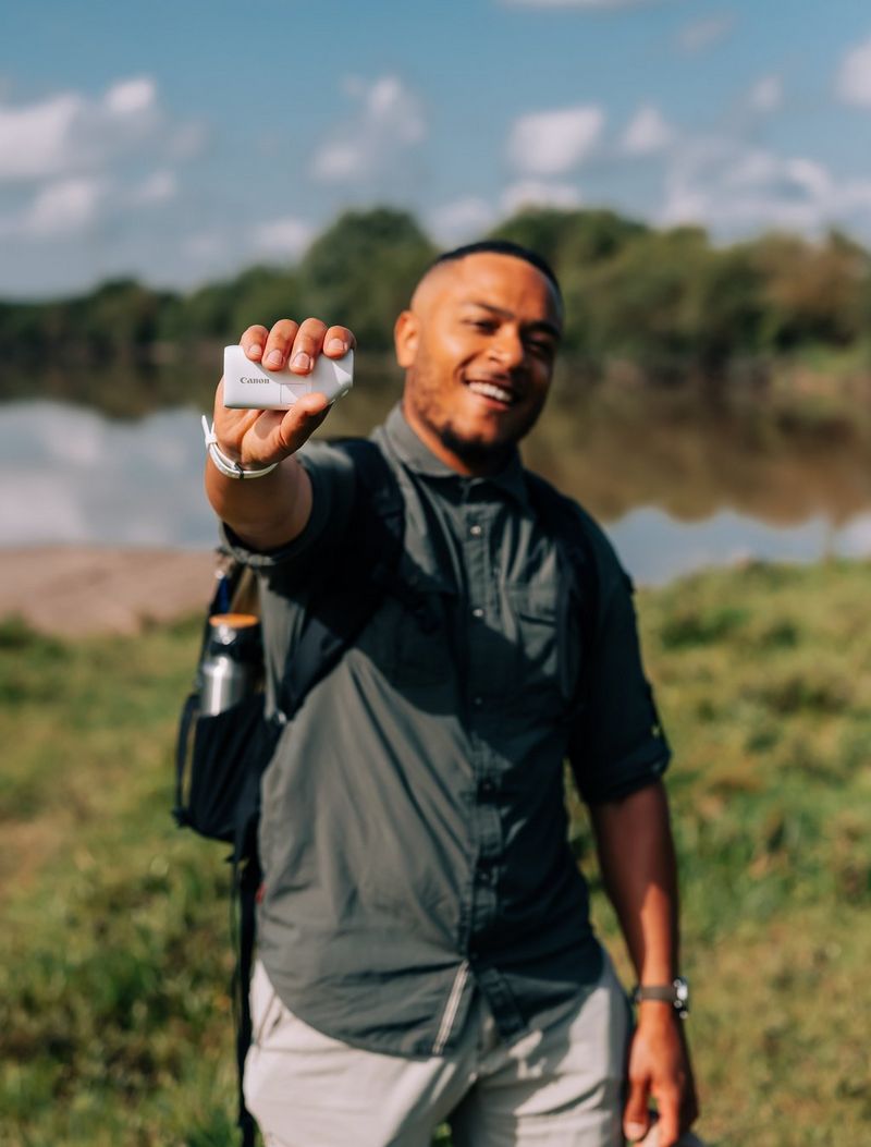 A man standing by a canal holding a Canon PowerShot ZOOM.