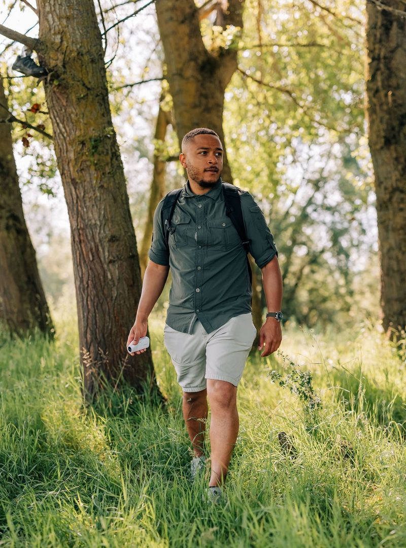 A man walking through trees in long grass, holding a PowerShot ZOOM in one hand.