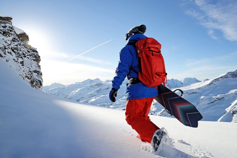 A snowboarder wearing blue and red winter clothing strides away from the viewer in a snowy mountainous landscape under a blue sky. © Richard Walch.